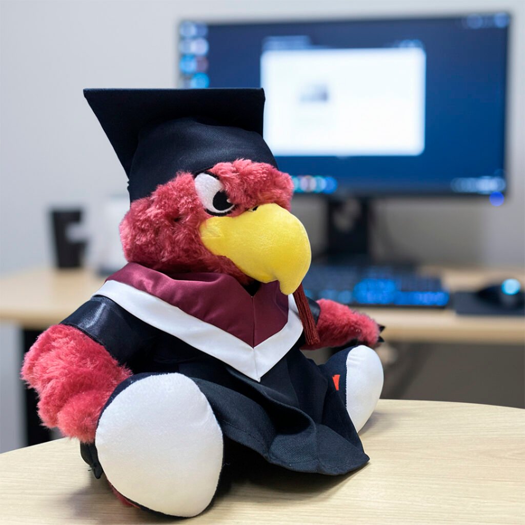 Peluche de Uvaldo (mascota aguila rapaz) sentado en una mesa de madera, vestido con toga y birrete de graduación en colores negro, blanco y borgoña (vino tinto). En el fondo, desenfocado, se ve un monitor de computadora en un escritorio de oficina. La mascota tiene plumaje rojo y un pico amarillo grande.