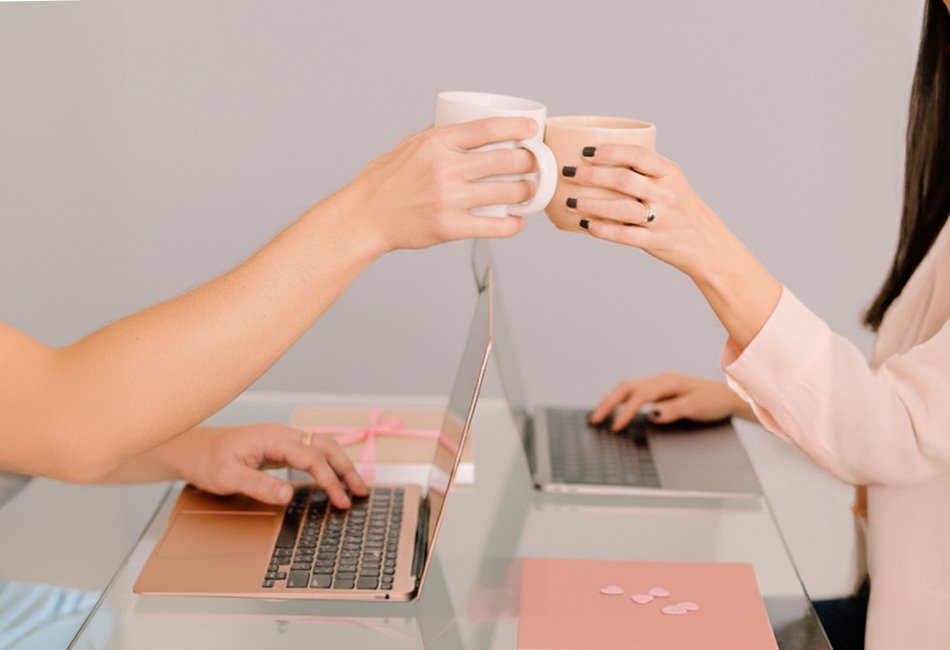 Dos personas frente a sus computadoras portátiles brindan con tazas de café sobre un escritorio, simbolizando una conexión saludable y respetuosa en un entorno virtual o académico.