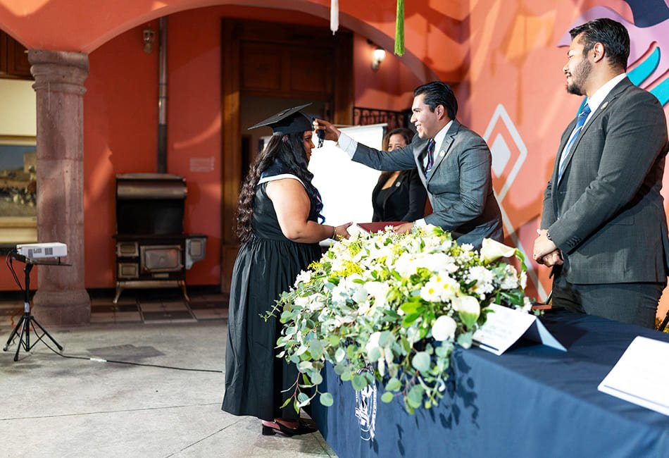 "Una estudiante de la Universidad Vizcaya en Línea recibe su diploma durante la ceremonia de graduación 2023-2026. Un directivo ajusta con orgullo la borla de su birrete frente a una mesa decorada con flores blancas, simbolizando la meta cumplida.
